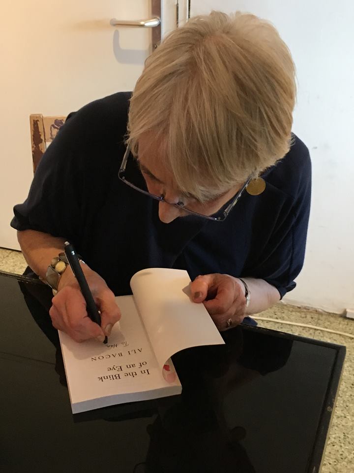 Inage shows Ali Bacon sated at a table signing a copy of her novel 'In the Blink of an Eye'