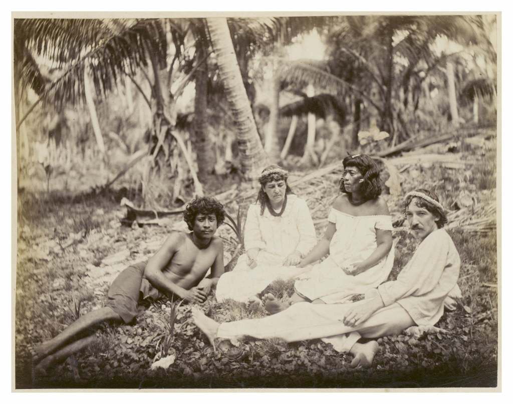Image is a sepia-toned photograph of four people sitting on the ground in a tropical forest with palm trees. Two women in white dresses (Fanny Stevenson on the left) sit in the middle, flanked by a shirtless young man on the left and a man in white clothing on the right (R. L. Stevenson)  who is reclining slightly. All four wear floral or woven headbands, suggesting a Polynesian or island setting. The group appears relaxed, seated among fallen leaves and tropical foliage.
