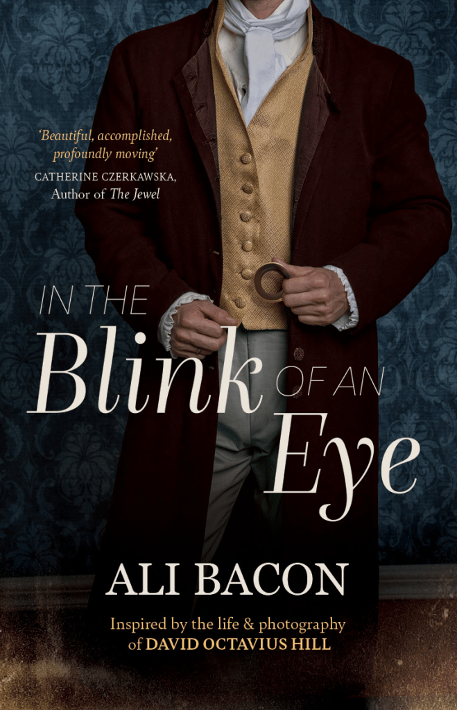 Book cover for "In the Blink of an Eye" by Ali Bacon. The image shows a man dressed in 19th-century clothing: a brown coat, yellow waistcoat, white cravat, and pale trousers. He holds a camera lens in one hand. The background features a dark blue patterned wallpaper. The cover includes a quote: “Beautiful, accomplished, profoundly moving” — Catherine Czerkawska, Author of The Jewel. The subtitle reads: “Inspired by the life & photography of David Octavius Hill.”