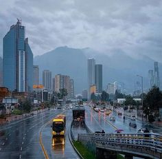 Image shows a cityscape with in the forgorund lanes of traffic, in the mid ground tall skyscrapers with mountain peaks shrouded in mist behind. 